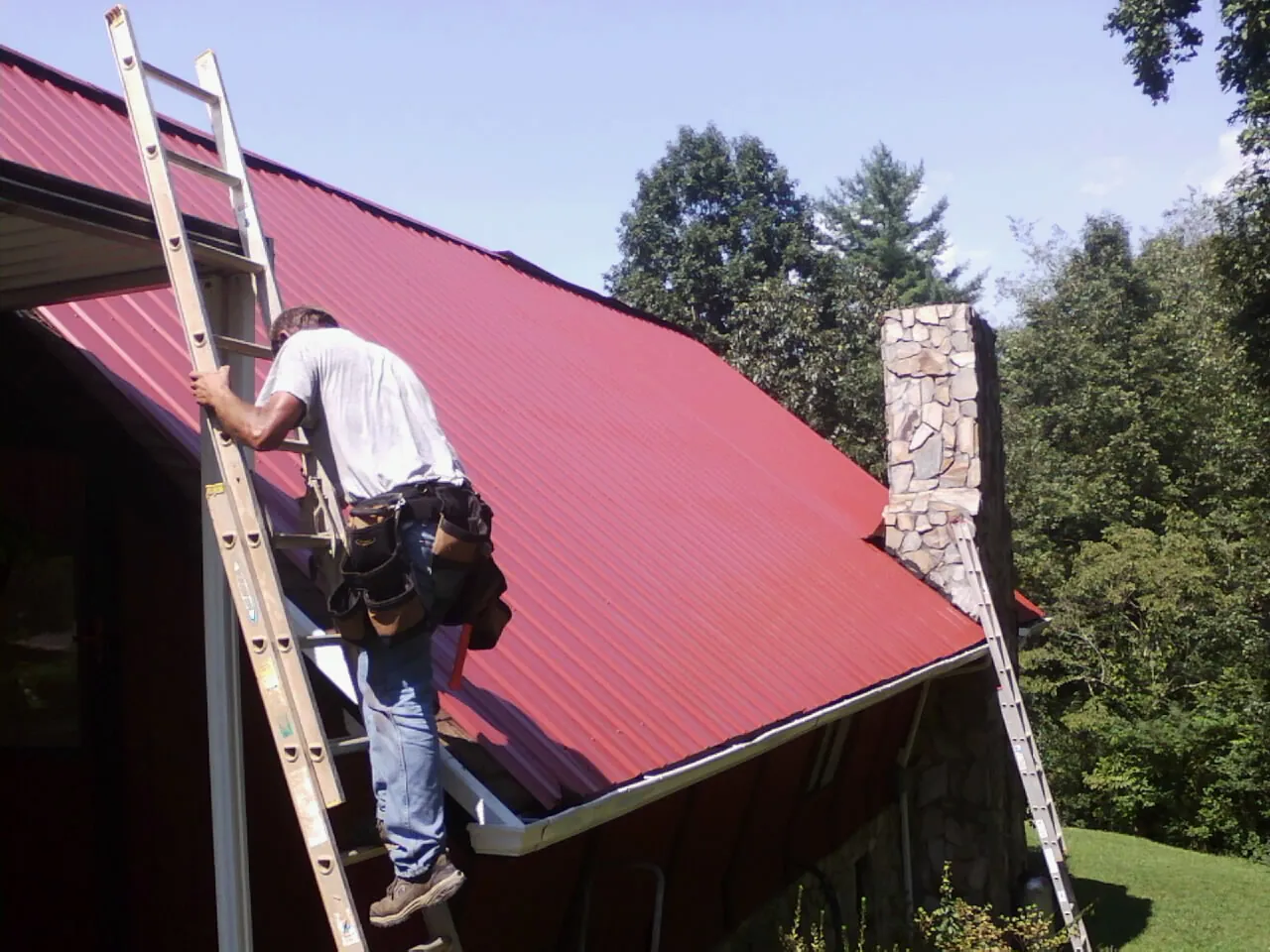 Roofer on a ladder beside a red metal roof and stone chimney — Soffit & Fascia Repair by Roofing Brothers in Preble