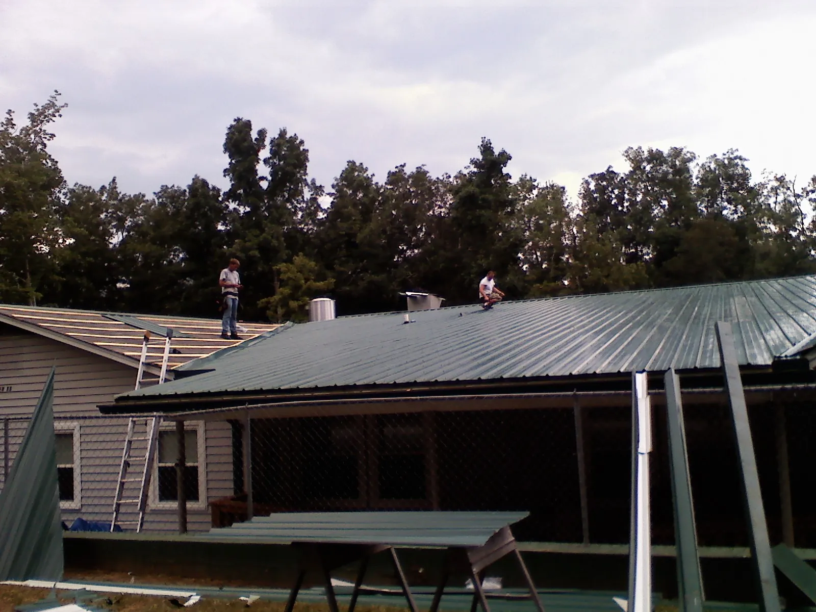Two roofers installing green metal panels above a covered porch in Preble